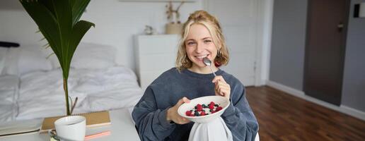 Portrait of smiling blond woman, eating breakfast, holding bowl and spoon, sitting in bedroom, looking happy at camera photo