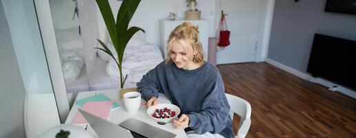 Portrait of smiling blond young woman, eating in front of laptop, watching s online while having breakfast, enjoying dessert, sitting in bedroom photo