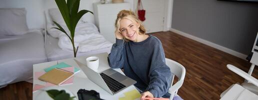 Portrait of smiling, beautiful blond woman, writing down notes, doing homework, studying from home, doing distance learning, online course, working remotely in her room photo