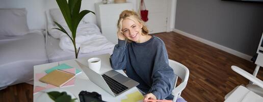 retrato de sonriente, hermosa rubio mujer, escritura abajo notas, haciendo tarea, estudiando desde hogar, haciendo distancia aprendiendo, en línea curso, trabajando remotamente en su habitación foto