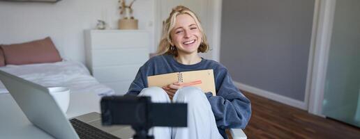 Portrait of smiling, charismatic young woman, content creator, records on digital camera, laughing and looking happy, using journal, writing in notebook photo