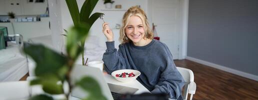 Portrait of young smiling woman, sitting in room, watching s on laptop, eating lunch or breakfast, healthy meal photo