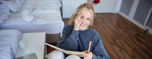 retrato de sonriente, carismático joven mujer, escritura abajo notas, haciendo planes y poniendo eso en planificador, participación diario, sentado en dormitorio y mirando contento a cámara foto