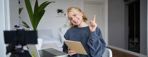 Portrait of young smiling, charismatic woman, raising one finger, teaching online, reading from notebook, looking at laptop, recording course, content maker working on new project from home photo