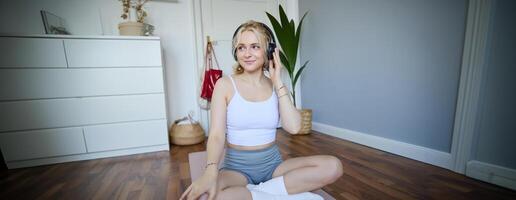 Portrait of happy, beautiful athletic woman, working out at home, listening music to boost energy while active training session, using wireless headphones and rubber yoga mat photo