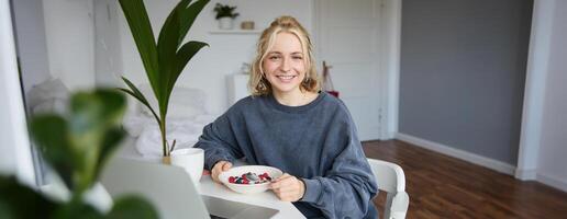 retrato de joven mujer comiendo sano comida en un habitación, acecho s en computadora portátil, teniendo almuerzo en frente de computadora, sonriente y mirando contento a cámara foto