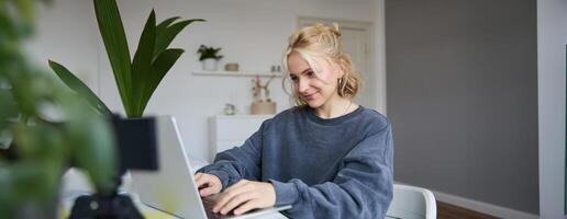 Portrait of young blond woman, female college student works from home on assignment, uses laptop, studies remotely, sits in room in front of computer photo