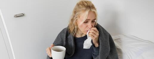 Portrait of woman catching a cold, staying home sick, drinking tea, sneezing in napkin, blowing nose, has covid symptoms photo