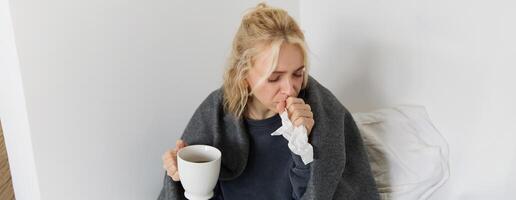 Concept of flu and quarantine. Close up portrait of young woman feeling sick, caught a cold, sneezing and blowing nose in napkin, holding warm tea in hand photo