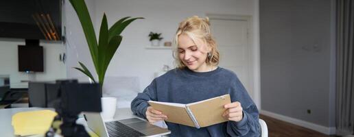 Portrait of young woman using digital camera and laptop, studying, connect to online live stream, records for vlog, creating content, holding notebook, sitting in room photo