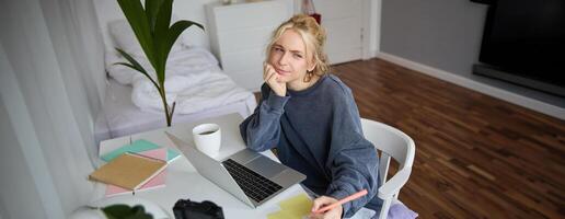 Portrait of young thoughtful girl, studying, making notes, writing down something in notebook, doing homework in her room, sitting in front of laptop, frowning while thinking photo