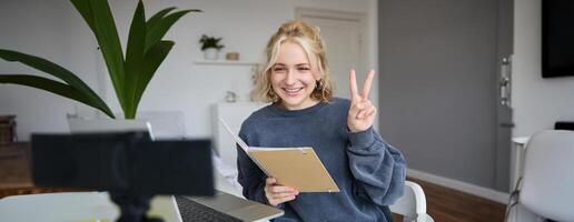 Image of smiling girl records of herself on digital camera, shows peace sign, sits in front of laptop, holds notebook photo