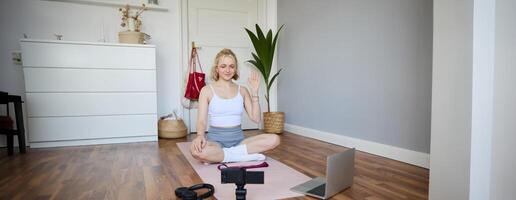 Portrait of young female athlete, fitness trainer recording vlog, training session on digital camera, sitting in a room on rubber yoga mat, showing exercises photo