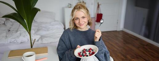 Portrait of cute smiling blond woman eats breakfast in her bedroom, looking at camera, holding bowl with dessert and spoon photo