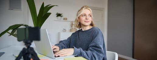 retrato de sonriente joven mujer, Universidad estudiante se sienta en su habitación, lo hace tarea, estudios remotamente desde hogar, usos ordenador portátil para Lanza libre trabajo foto