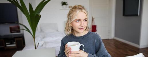 Close up portrait of smiling young blond woman, sits with cup of tea in bedroom, rests at home, enjoys her coffee photo