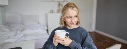 Portrait of smiling beautiful young woman, sitting in bedroom with cup of tea, resting at home alone, enjoys her weekend indoors photo