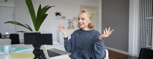 Portrait of young social media content creator, woman sits in room with laptop and digital camera, records lifestyle vlog, says hello, chats to someone online via computer photo