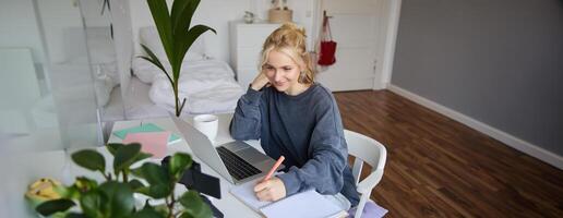 retrato de sonriente, hermosa rubio mujer, escritura abajo notas, haciendo tarea, estudiando desde hogar, haciendo distancia aprendiendo, en línea curso, trabajando remotamente en su habitación foto