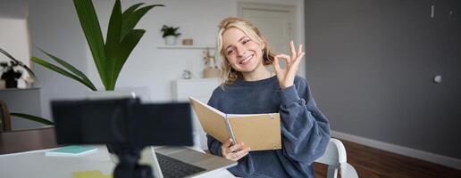 Portrait of cute, stylish young woman, shows okay sign, holds notebook, reads aloud her notes, records vlog, looks at digital camera and makes ok gesture photo