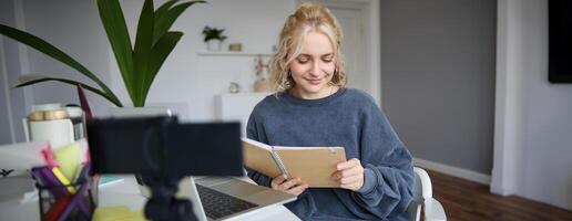 Portrait of young woman with notes, working from home, recording on digital camera, live streaming from her room, reading from notebook and smiling photo