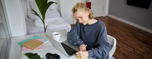 retrato de sonriente joven mujer, hembra alumno, haciendo distancia aprendizaje curso, utilizando computadora portátil, estudiando a hogar, escritura abajo, haciendo notas foto