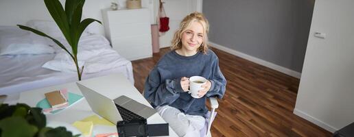 imagen de joven mujer, social medios de comunicación hombre de influencia, edición su en computadora portátil, se sienta en un habitación con computadora y digital cámara, Bebiendo café, sonriente a cámara foto