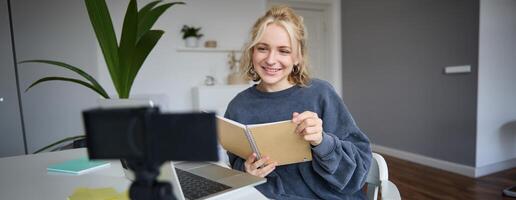 Portrait of young woman using digital camera and laptop, studying, connect to online live stream, records for vlog, creating content, holding notebook, sitting in room photo