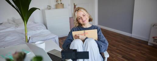 Portrait of blond smiling woman, records on digital camera how she writes in notebook, talks to followers, doing lifestyle blog content in her room photo