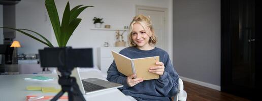 Young smiling blond woman, sits near laptop, uses digital camera to record blog, creates lifestyle content for social media, records tutorial, shows notebook, reads her notes photo