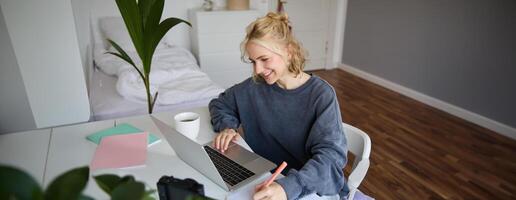 retrato de sonriente joven mujer, hembra alumno, haciendo distancia aprendizaje curso, utilizando computadora portátil, estudiando a hogar, escritura abajo, haciendo notas foto