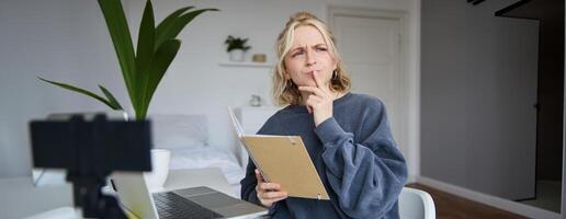 retrato de joven mujer, estudiante sentado en habitación, estudiando desde hogar, remoto educación concepto, participación cuaderno y pensamiento, mirando aparte con pensativo rostro, concentrando en asignación foto