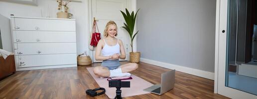 Portrait of young fitness blogger, woman showing exercises to her followers, recording on digital camera, doing workout training session, home yoga, sitting in front of laptop on rubber mat photo