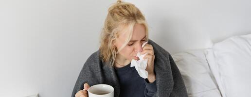 Concept of flu and quarantine. Close up portrait of young woman feeling sick, caught a cold, sneezing and blowing nose in napkin, holding warm tea in hand photo