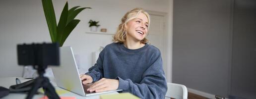 Portrait of young blond woman, female college student works from home on assignment, uses laptop, studies remotely, sits in room in front of computer photo