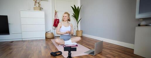 Portrait of young fitness blogger, woman showing exercises to her followers, recording on digital camera, doing workout training session, home yoga, sitting in front of laptop on rubber mat photo