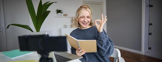 Portrait of cute blond girl with notebook in hands, shows okay sign, records , social media content, blogging from her room photo