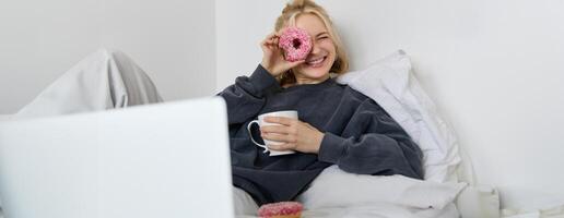 Portrait of happy cute woman, lying in bed with laptop, drinking tea, looking through doughnut hole and smiling at camera photo