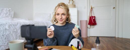 Portrait of young beauty blogger, woman gives advices to audience, how to apply makeup, sits in front of digital camera, holds foundation cream and cotton pad photo