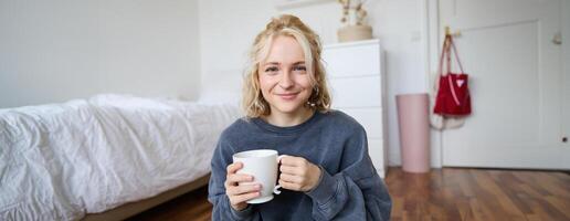 Image of young teenage girl sitting in her bedroom on floor, drinking cup of tea and enjoying day at home, smiling and looking at camera photo