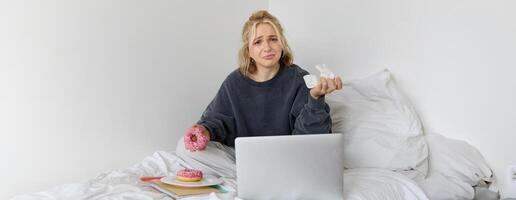 Portrait of disappointed, depressed young woman, crying, sitting on bed with laptop, eating comfort food, holding doughnut and wiping tears off photo