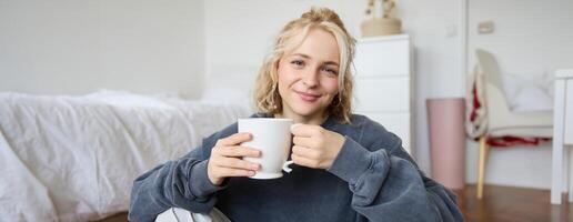 retrato de joven mujer sentado en dormitorio piso, Bebiendo té, participación blanco jarra y sonriente a cámara foto