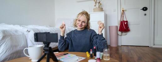 Image of young woman, vlogger records on digital camera, shows beauty products, recommending makeup for audience on social media, sits on front in a room photo