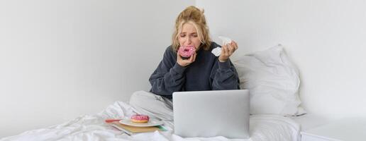 retrato de triste, llorando joven mujer, quedarse a hogar, sentado en cama con rosquilla y comodidad alimento, mirando a alguna cosa molesto en ordenador portátil pantalla foto