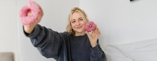 Portrait of beautiful smiling blond woman, showing two pink doughnuts at camera, eating delicious food photo