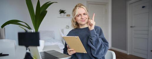 Portrait of young creative woman, content maker, sitting in room, working from home, using laptop, holding notebook, raising finger, has an idea, eureka gesture photo