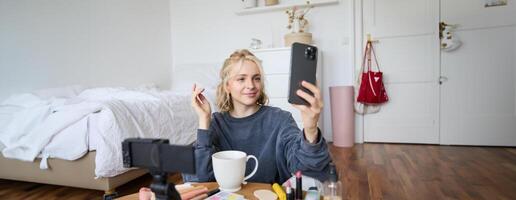 Portrait of young beauty blogger, recording on digital camera, doing online live stream while applying makeup, talking to her followers, sitting on bedroom floor photo