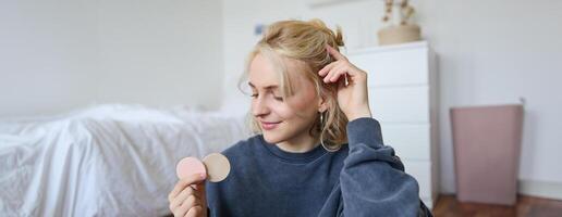 Portrait of young woman chatting on live stream about makeup, sits on floor in bedroom, showing beauty products to followers photo