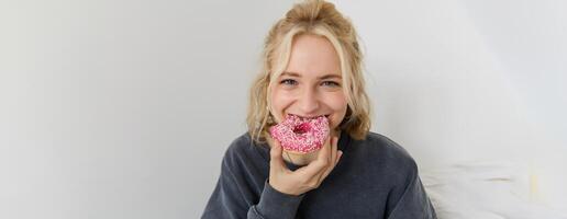 Portrait of cute blond girl holding pink doughnut with sprinkles on top, showing her favourite food photo