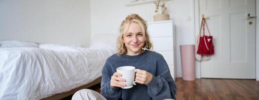 retrato de joven mujer sentado en dormitorio piso, Bebiendo té, participación blanco jarra y sonriente a cámara foto